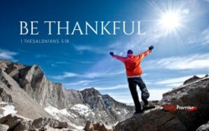 A man standing in the mountains with hands raised - "Be Thankful"