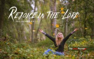 A woman is sitting in the woods with her hands raised in joy. "Rejoice in the Lord" - Philippians 4:4