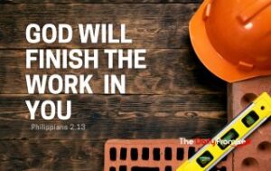 Several construction related items like a hard hat, bricks and a level laying on a dark wood background. "God Will Finish the Work in You"
